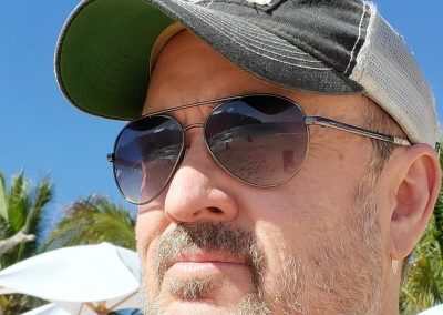 Man wearing sunglasses and a distressed baseball cap outdoors on a sunny day with parasols in the background.